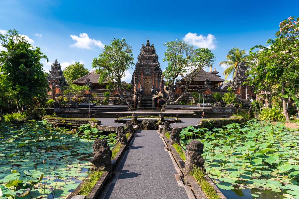 A view of the Saraswati Temple in Ubud, Bali. The temple is surrounded by greenery, lotus ponds, and Balinese architecture under a bright blue sky. The path leading to the temple is flanked by intricately carved stone statues.