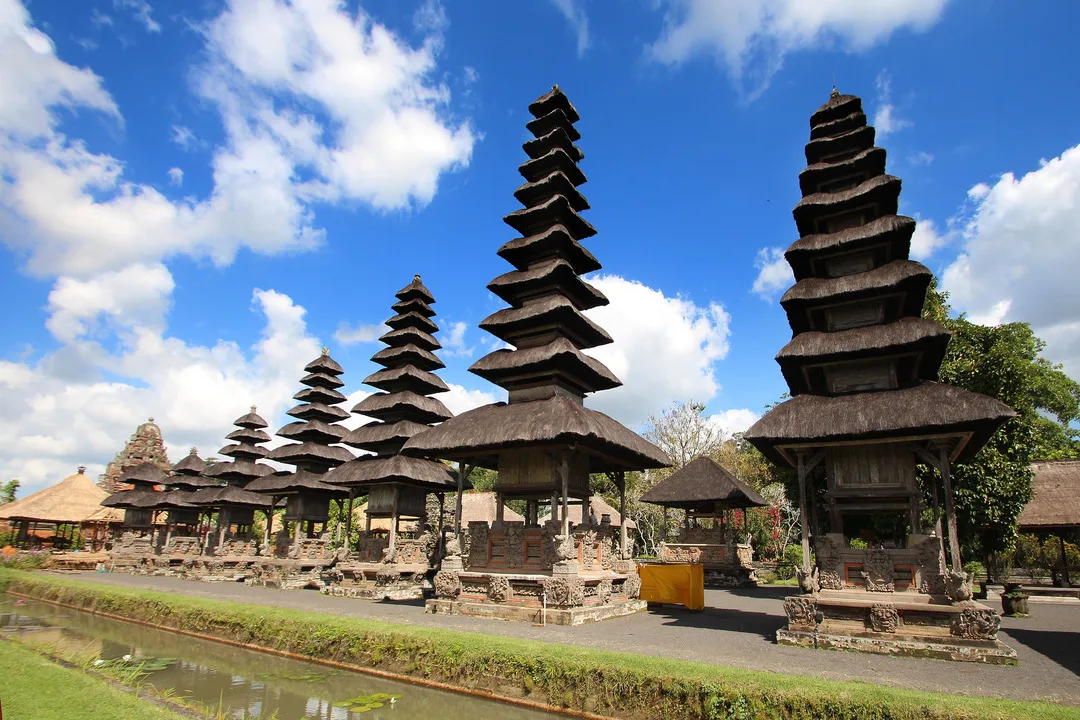 The Taman Ayun Temple in Bali features a multi-tiered pagoda-like shrine with traditional thatched roofs. The temple is surrounded by greenery, above is a clear blue sky with scattered clouds, and a reflective canal is in the foreground. 