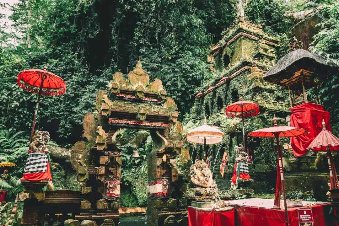 Taman Pecampuhan Sala Temple in Bali, Indonesia with greenery surrounding it. The temple features moss-covered stone structures, red umbrellas and intricate carvings, including statues of deities.