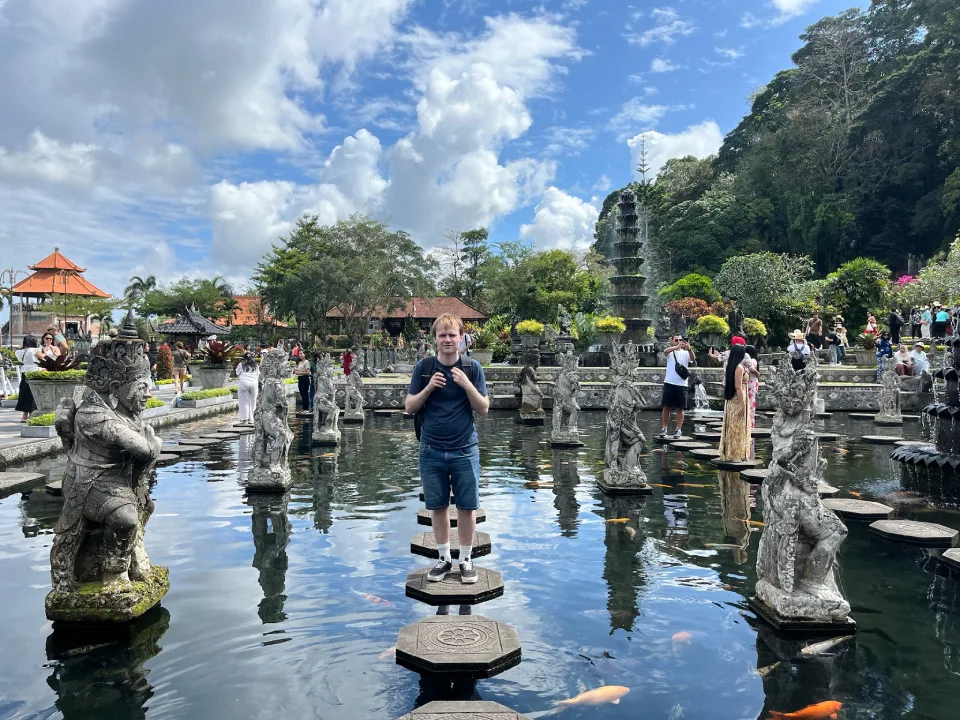 A person standing on stepping stones in the middle of the main pond at Tirta Gangga Water Palace in Bali, Indonesia. The pond features orange koi fish, stone statues of deities, and greenery, with temple structures and visitors in the background under a bright, partly cloudy sky. 