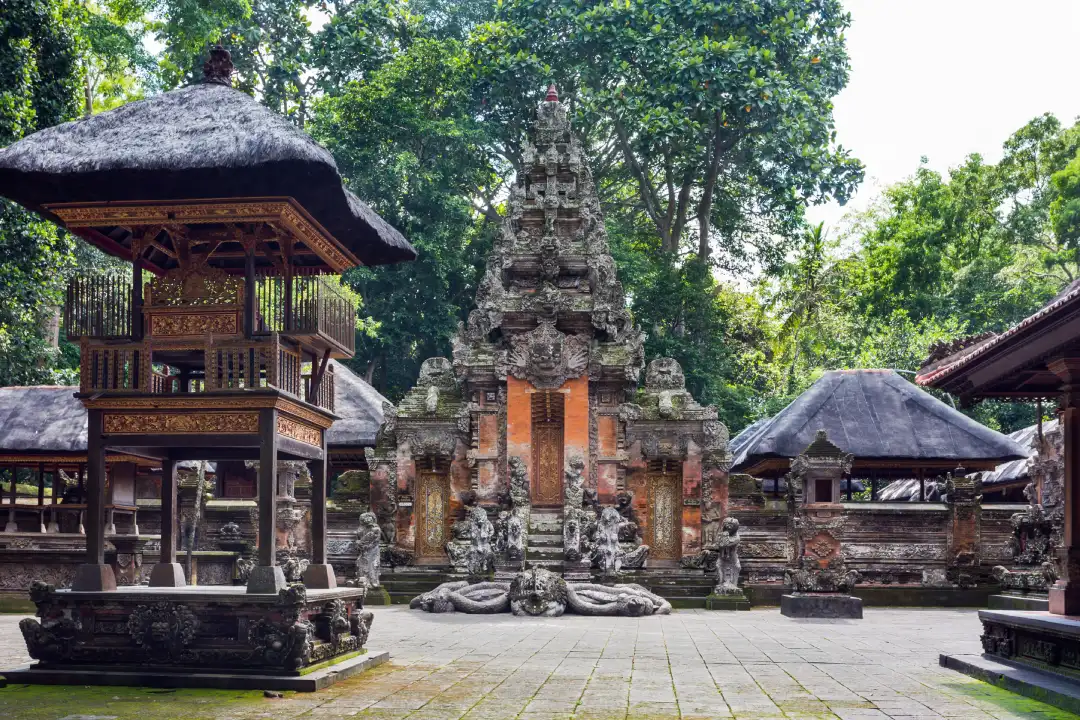 A Balinese temple in Ubud Sacred Monkey Forest. You can see intricately carved stone structures, a thatched roof pavilion, and a green forest in the background.