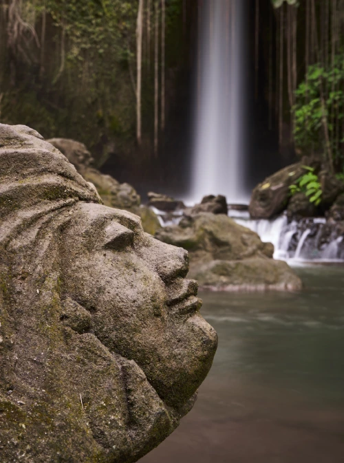 A detailed stone carving of a human face on a rock, positioned near the edge of a waterfall. The background features the cascading waters of Sumampan Waterfall, surrounded by green foliage and moss-covered stones.