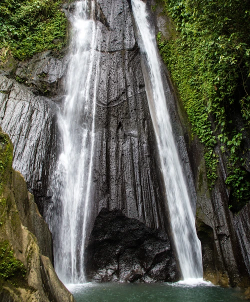 Dusun Kuning Waterfall with two streams of water cascading down dark, wet rock faces into a clear pool below. The surrounding cliffs are covered in green vegetation, creating a natural backdrop. 