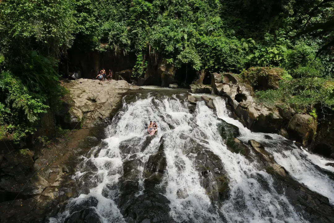 Goa Rang Reng Waterfall near Ubud, Bali, is surrounded by green vegetation and rocky terrain. Two people are sliding down the cascading water in the foreground, while others are visible sitting on the rocks nearby, enjoying the natural setting. 