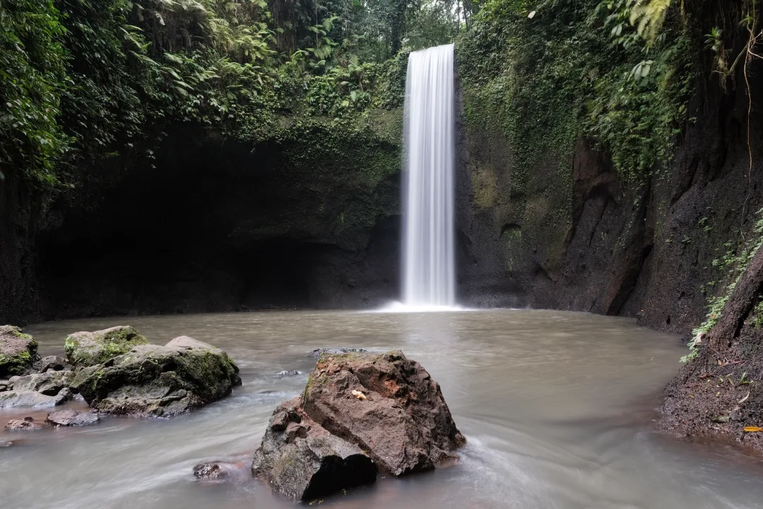 Waterfall in a tropical setting, with water cascading into a calm pool surrounded by moss-covered rocks and dense greenery.