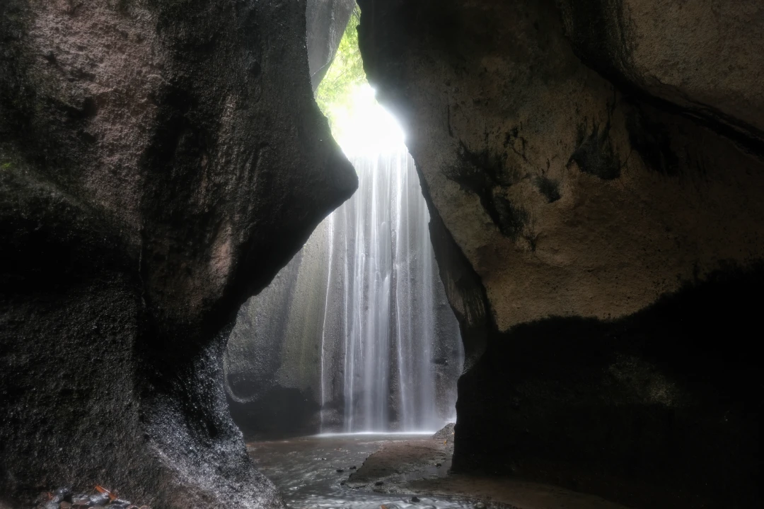 A view of Tukad Cepung Waterfall in Bali, framed by towering rock walls and illuminated by soft sunlight rays filtering through the opening above. The waterfall cascades gently into a small pool, creating a serene, hidden oasis.