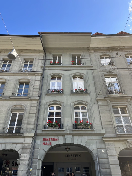 Facade of the Einstein House in Bern, Switzerland, a grey historic building with an arched entryway labelled “Einstein” and red flowers on window sills. A sign reading “Einstein Haus” is painted in red beside the arch