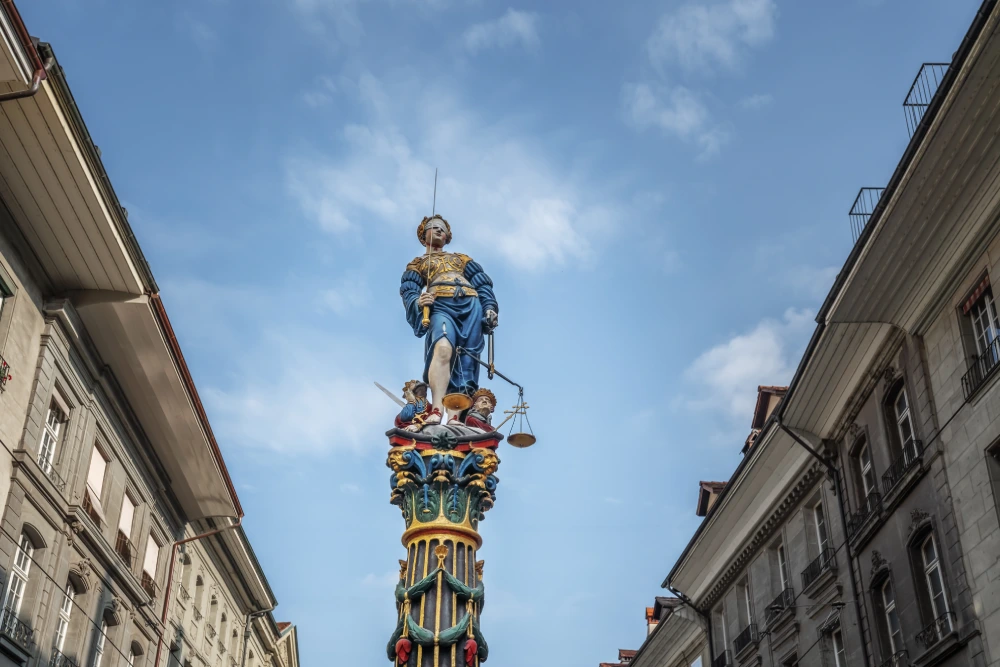 Statue of Lady Justice in Bern, Switzerland, standing atop a fountain, holding scales and a sword against a bright sky between buildings.