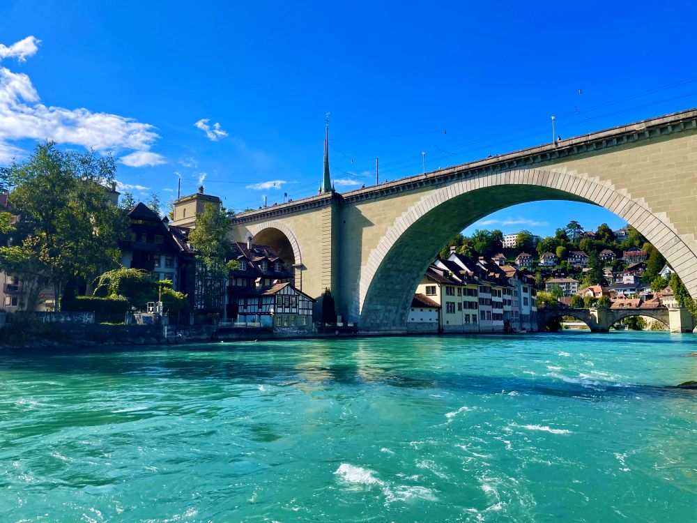 Sunny day in Bern, Switzerland, with the Nydeggbrücke bridge crossing over the bright blue Aare River, surrounded by old houses and green hills.