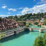View of old buildings with red roofs along a turquoise river in Bern, Switzerland, with a stone bridge and church tower under a blue sky.