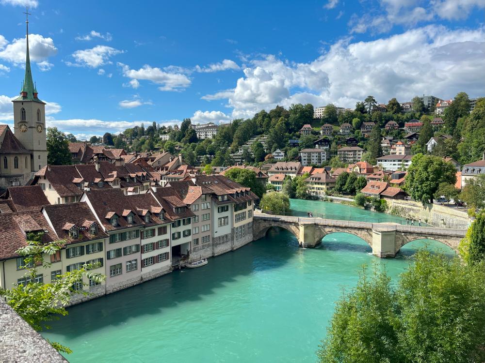 View of Bern's red-roofed medieval buildings along the Aare River on the Nydeggbrücke stone bridge and green hills nearby, all under a bright blue sky with scattered clouds.