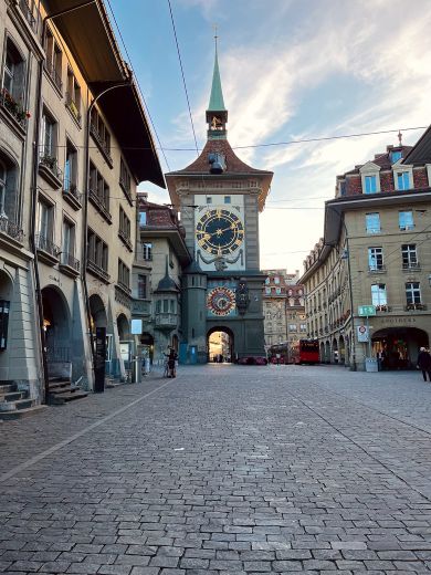 View of the Zytglogge clock tower in Bern, featuring a large astronomical dial and medieval stone archway, surrounded by historic buildings along a wide cobblestone street under a lightly clouded sky.