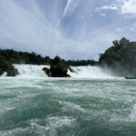 A long-distance view of Rhine Falls from the river. A tour boat can be seen approaching the falls on the right in the distance, and a Laufen Castle on a wooded hill above.