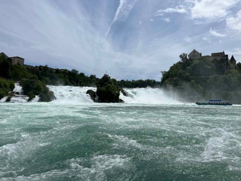 A long-distance view of Rhine Falls from the river. A tour boat can be seen approaching the falls on the right in the distance, and a Laufen Castle on a wooded hill above.
