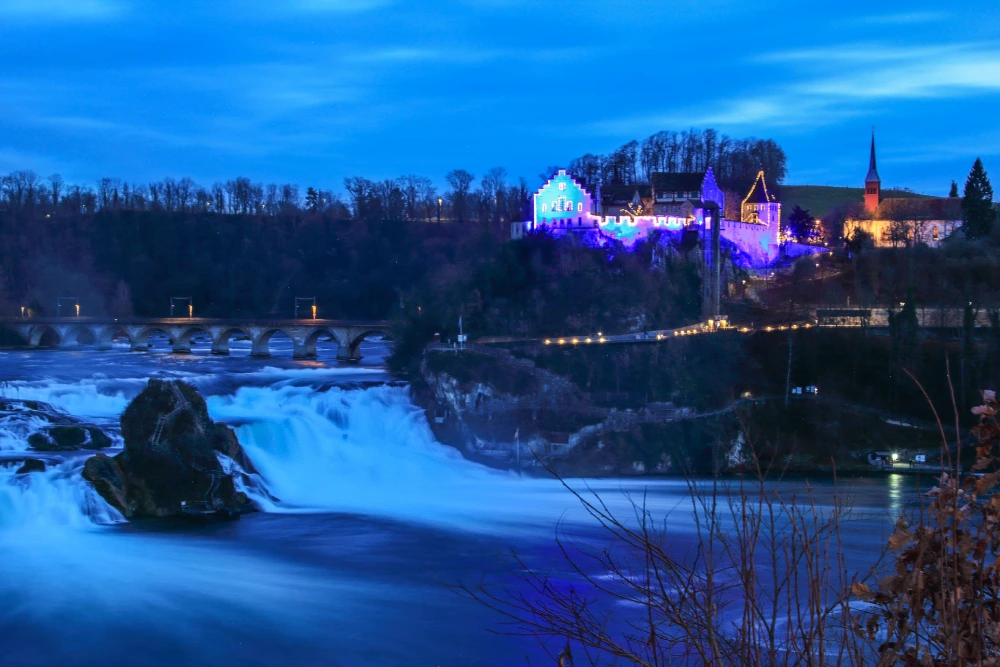 Nighttime view of Schloss Laufen Castle illuminated with blue and purple Christmas lights above the Rhine Falls in Switzerland.