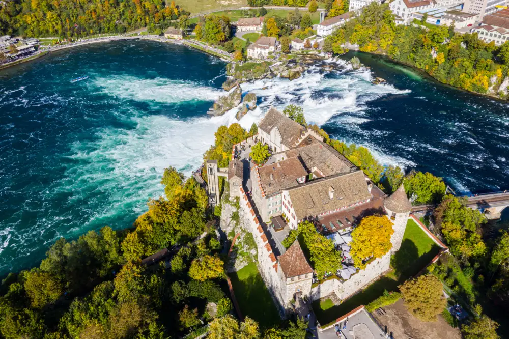 Aerial view of Schloss Laufen overlooking the Rhine Falls, with autumn trees and turquoise water below.
