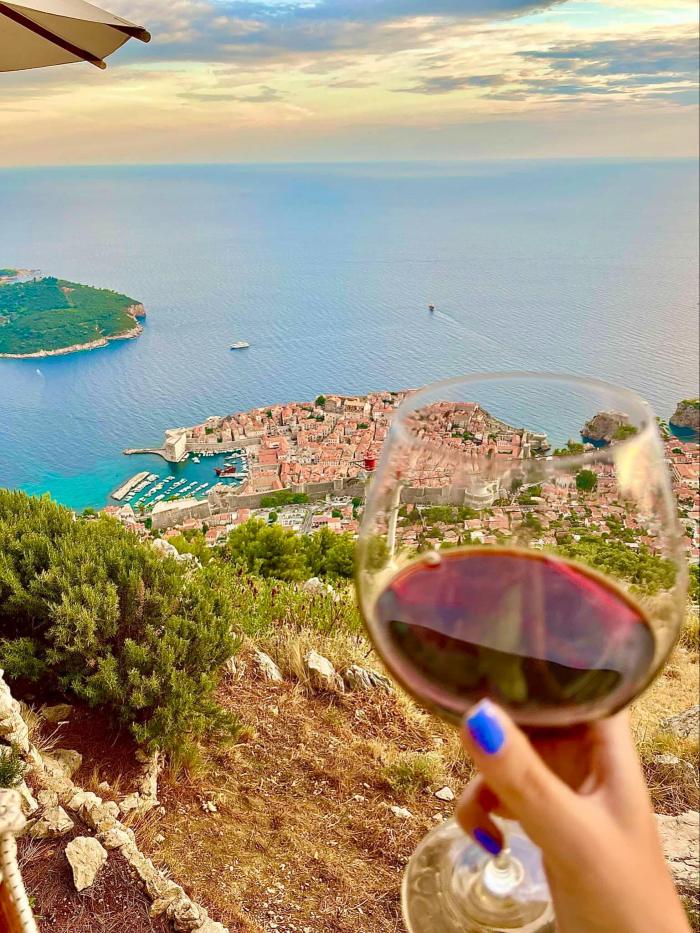 A glass of red wine in the foreground, with a scenic view of Dubrovnik's old town and the Adriatic Sea in the background. 