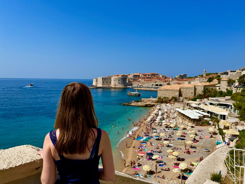 A woman overlooking Banje Beach in Dubrovnik, Croatia. In the background is a busy beach with umbrellas and sunbathers, and Dubrovnik’s Old Town walls.